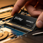 A close-up, high-angle shot of a person's hand carefully installing a black Kingston FURY Impact SODIMM RAM module into the memory slot of an open laptop motherboard, with a precision screwdriver resting nearby on a dark workspace.